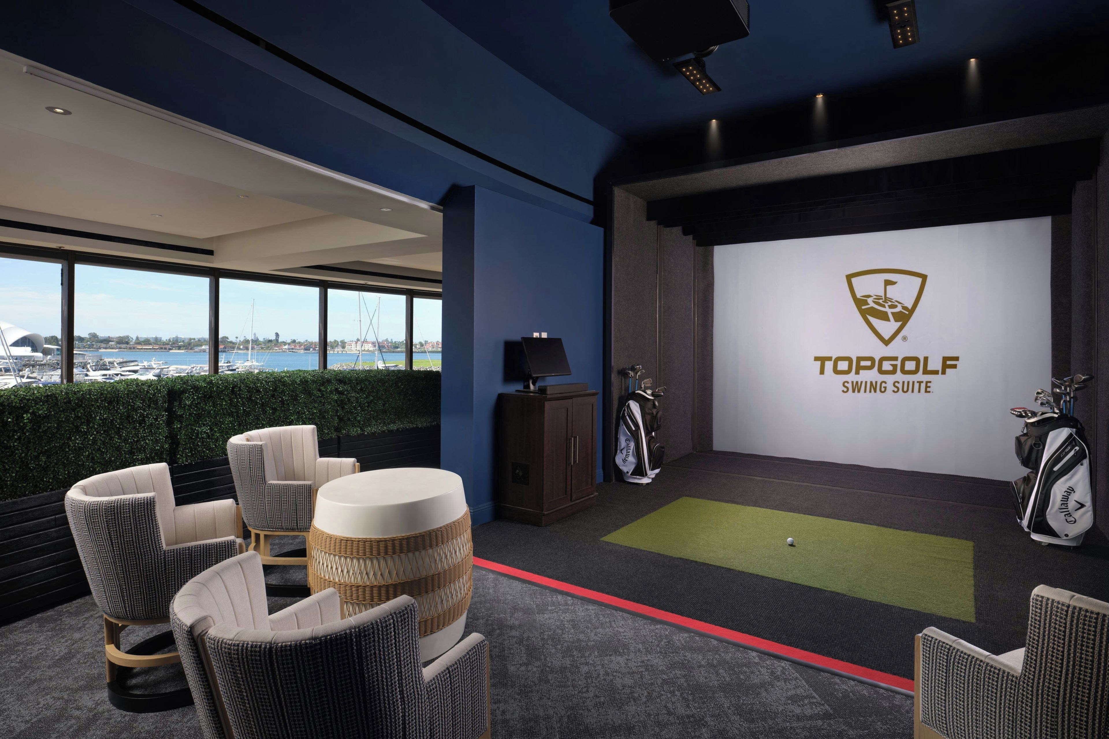A modern indoor golf simulation room with a view of the marina, comfortable seating, and a Targotle swing suite display.