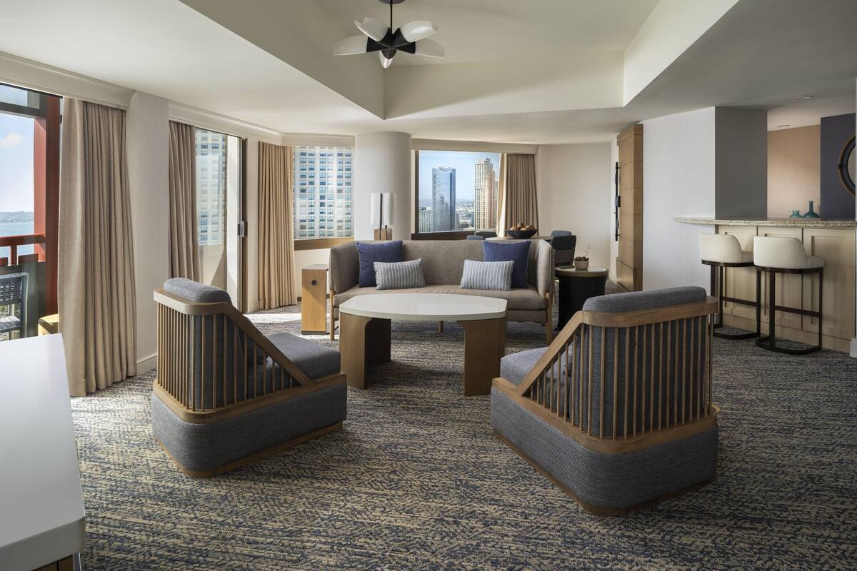 A modern hotel room with a large window, a round coffee table, two armchairs, a sofa, a small kitchen area, and a balcony.