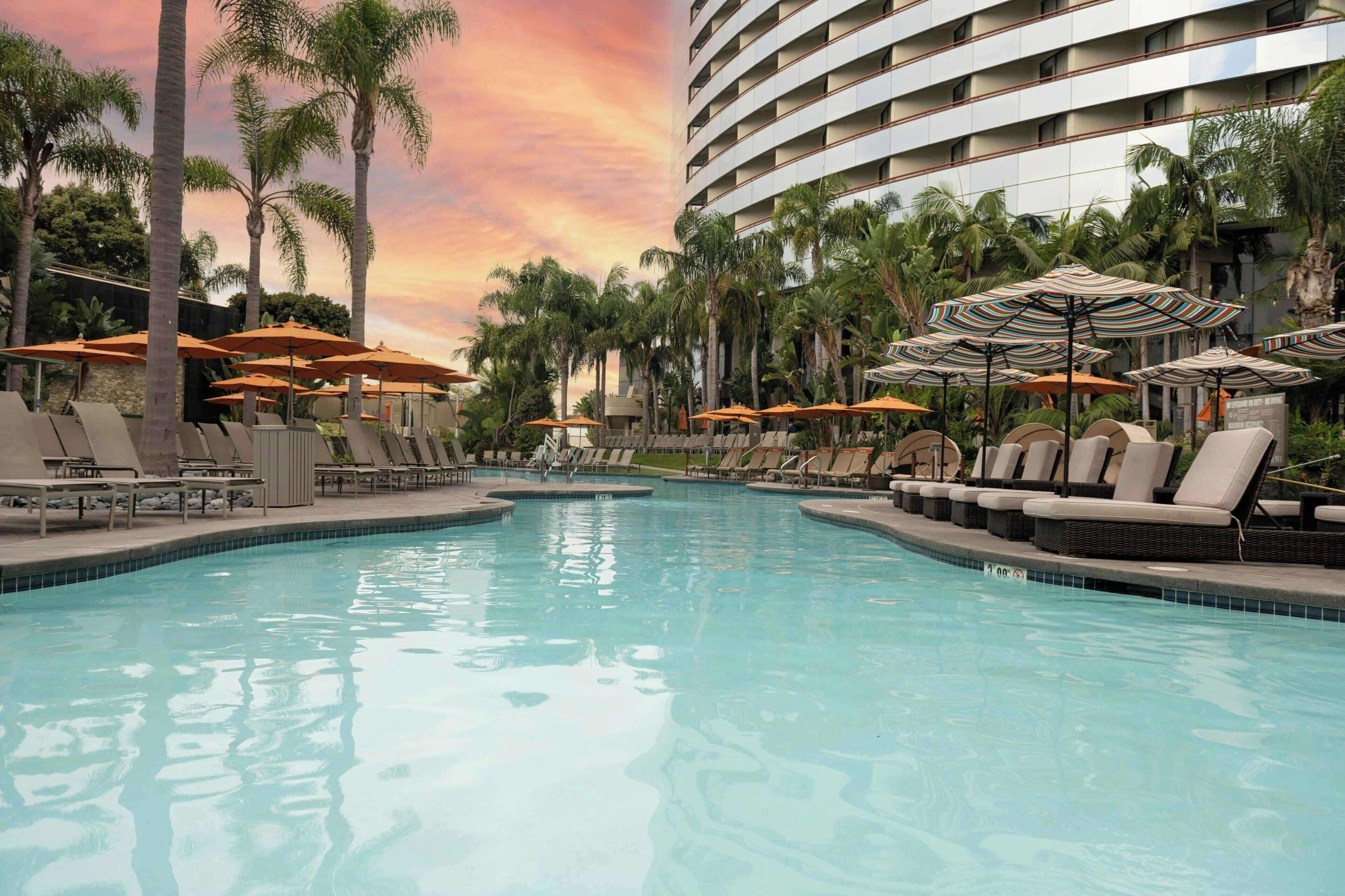 A swimming pool with lounge chairs and umbrellas, surrounded by palm trees and a hotel building.