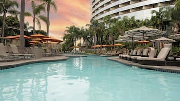 A swimming pool with lounge chairs and umbrellas, surrounded by palm trees and a hotel building.