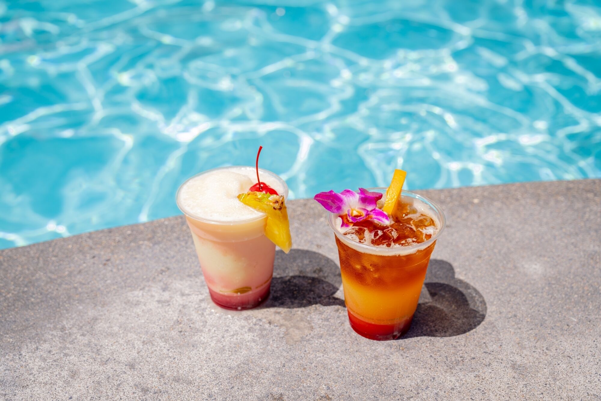 Two drinks with fruit garnishes on a stone surface by a pool.