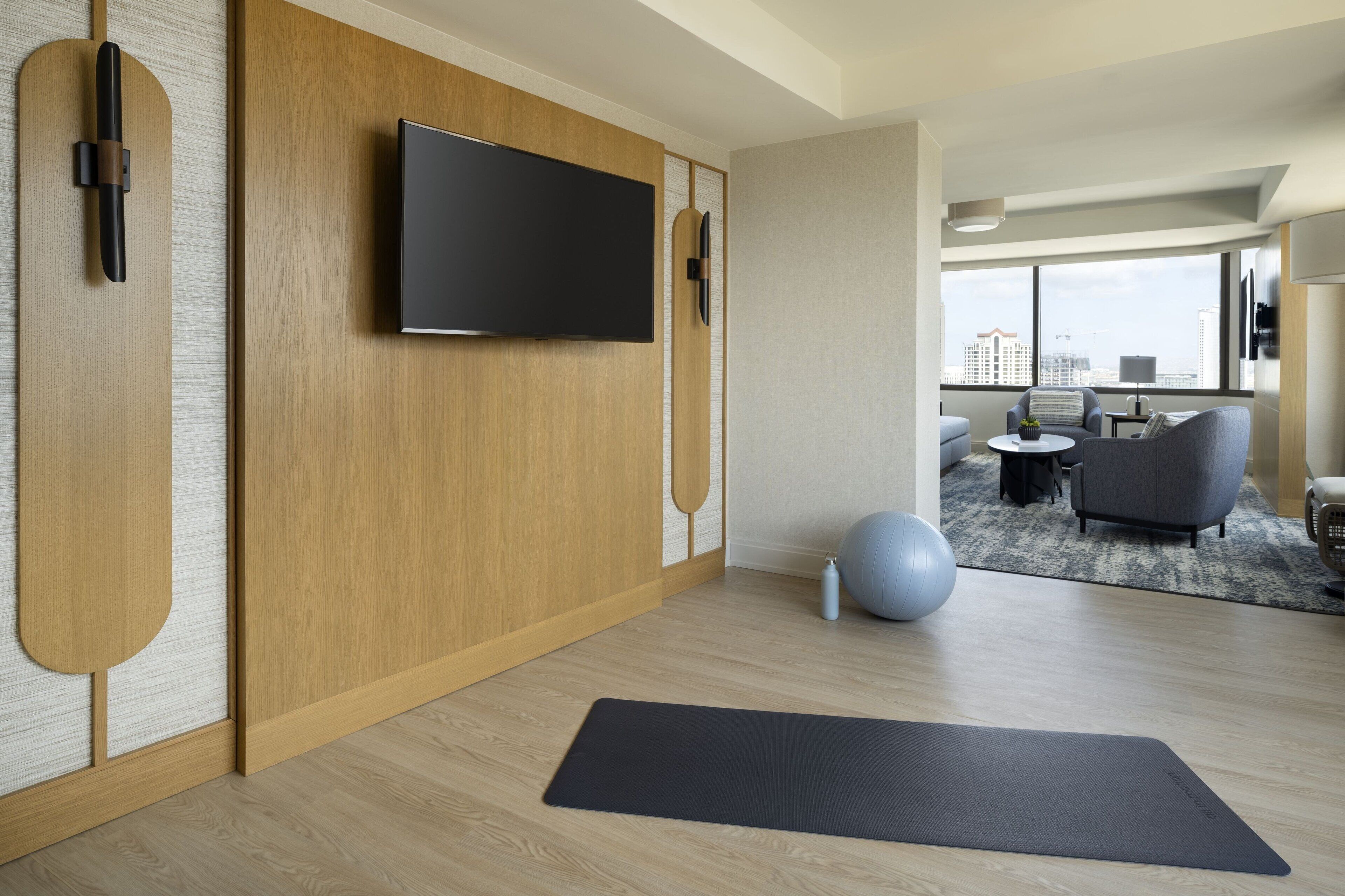 A modern hotel room with a wooden wall panel, a mounted TV, a yoga mat, a blue exercise ball, and a city view through large windows.