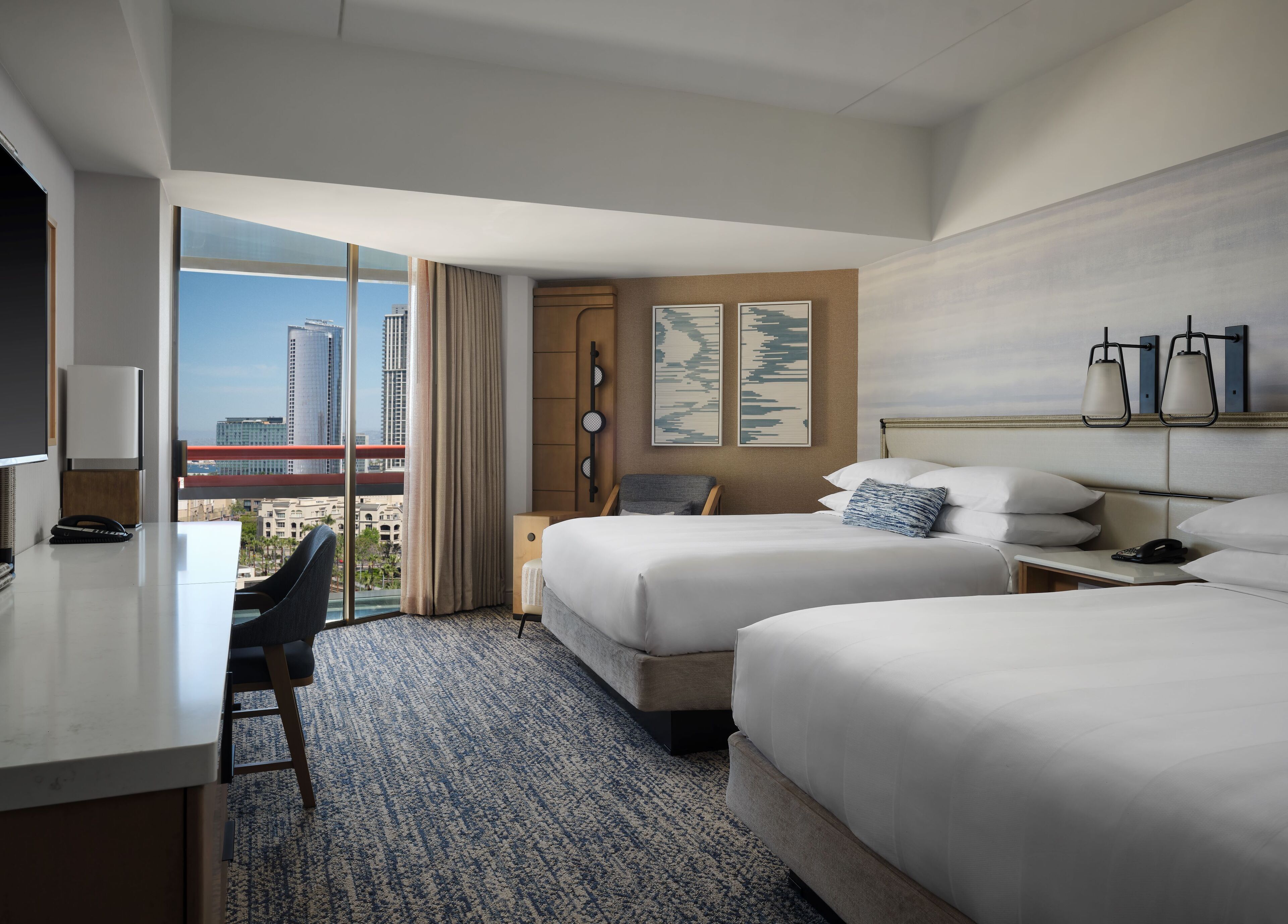 A hotel room with two beds, a desk, and a chair. There is a large window with a view of buildings and a red bridge.