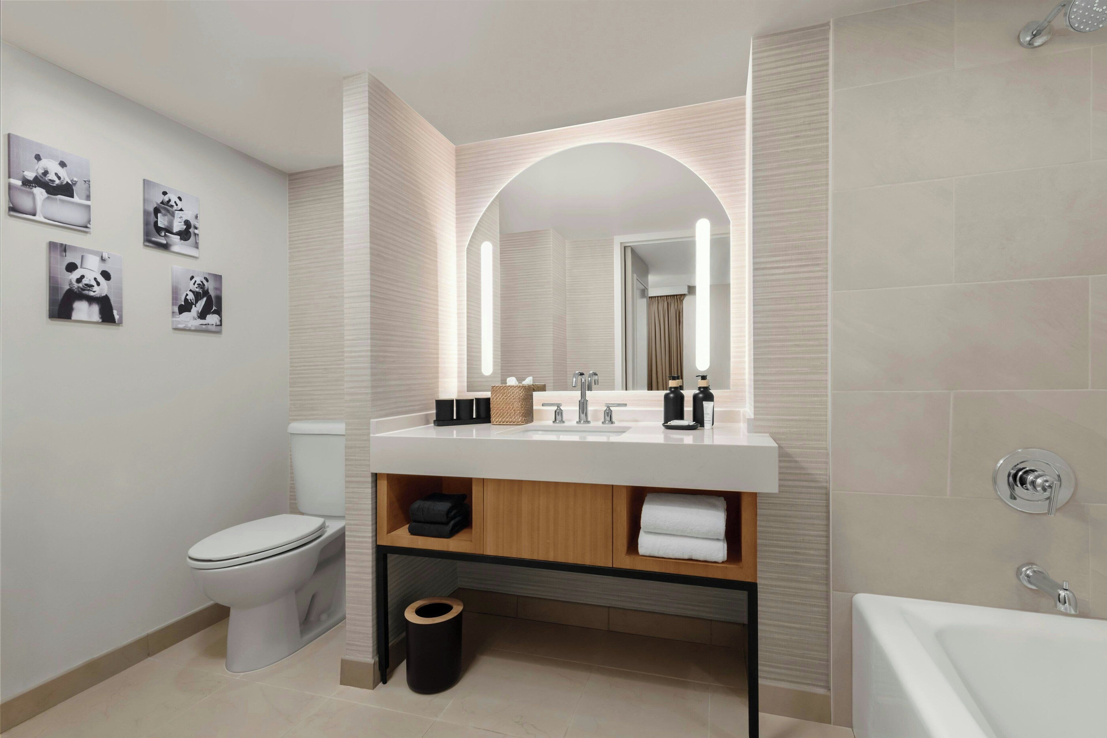 Een moderne badkamer met toilet, wastafel en bad. Er hangt panda-thema decoratie aan de muur en een spiegel met ingebouwde verlichting.