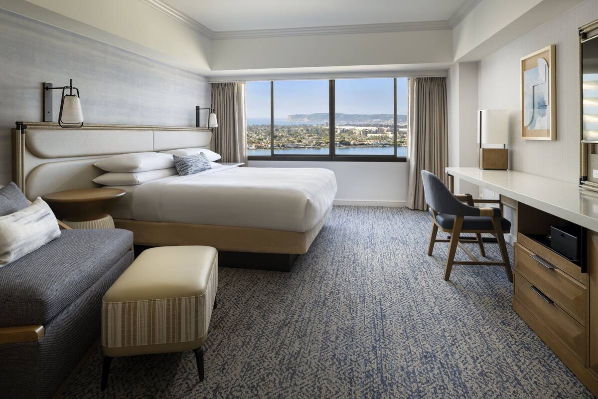 A hotel room with a large bed, a desk, a chair, and a view of a cityscape through a large window.