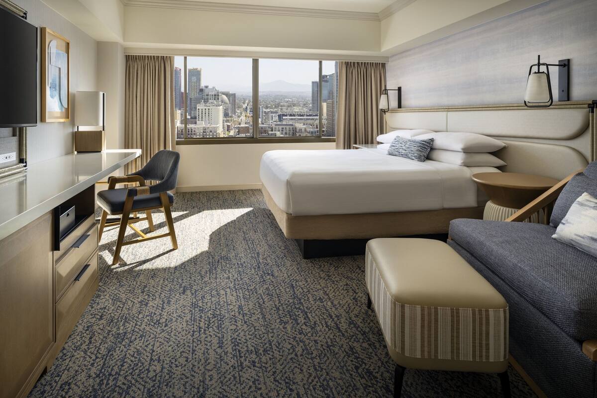 A hotel room with a large bed, a desk, a chair, a sofa, and a city view through the window.