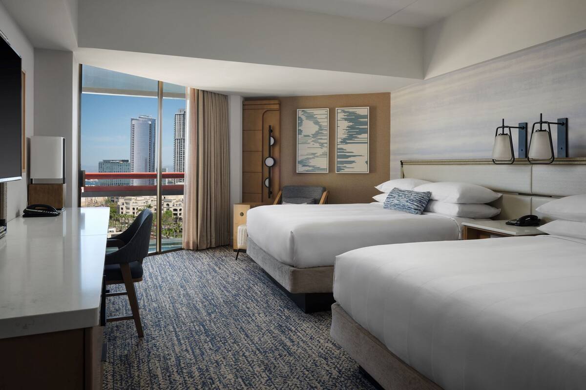 A hotel room with two beds, a desk, and a large window offering a city view.