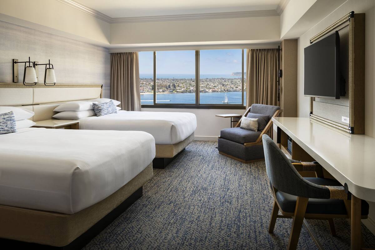 A hotel room with two beds, a desk, a chair, a TV, and a large window with a city view.