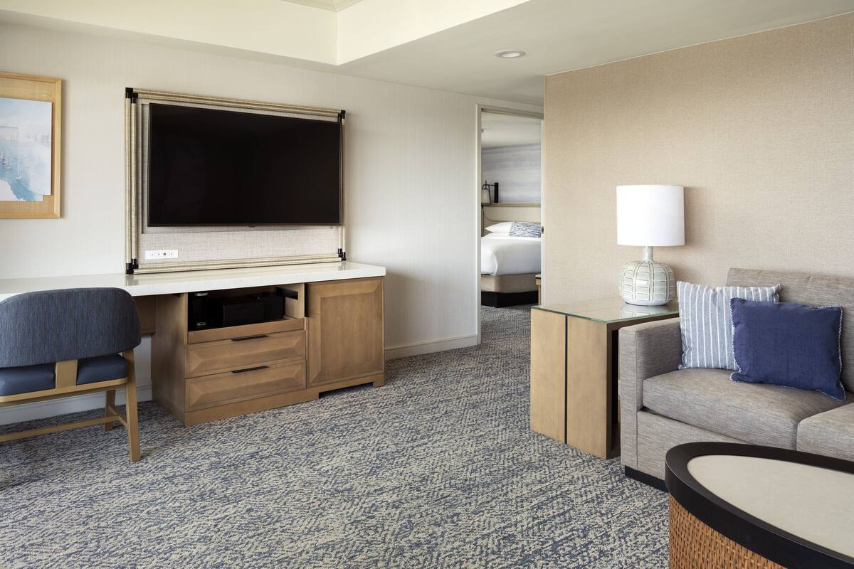 A hotel room with a large flat-screen TV, a desk with drawers, a comfortable sofa, a small table with a lamp, and a bed in the background.