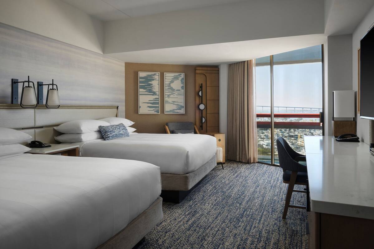 A hotel room with two beds, a desk, a chair, a large window with a city view, and a clock on the wall.