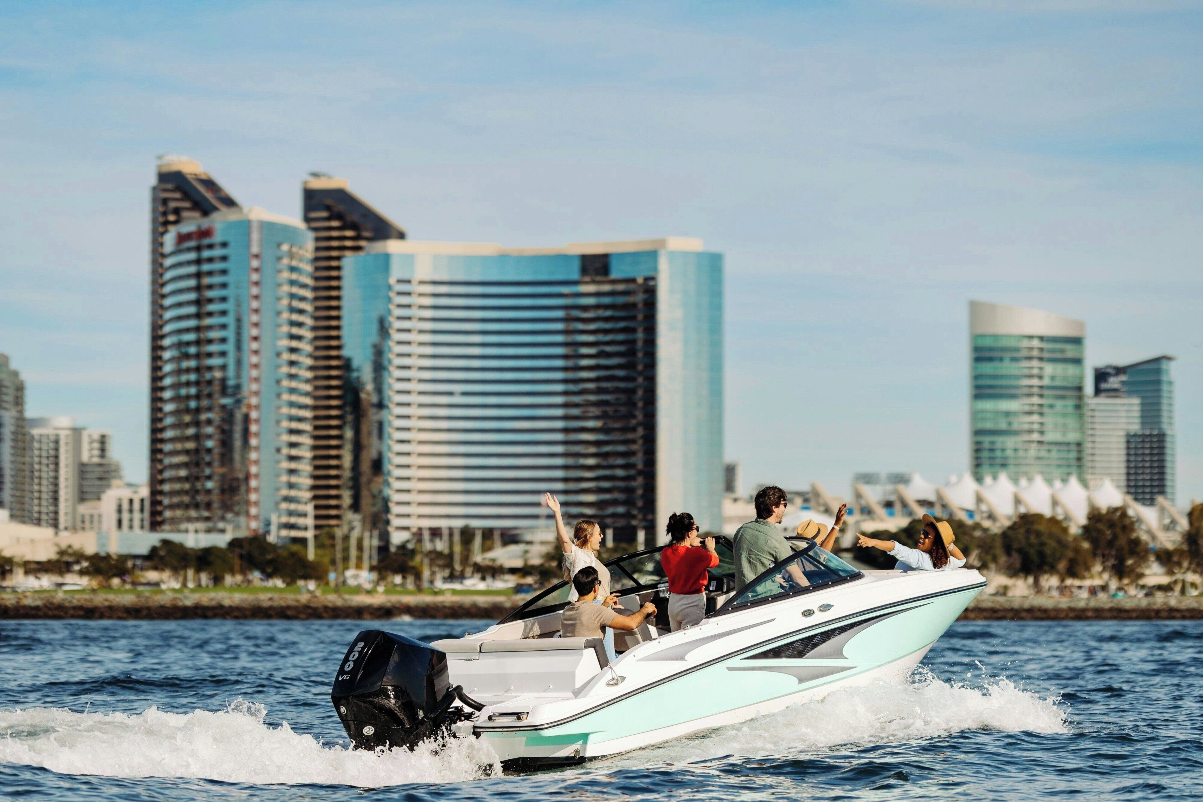 A speedboat with passengers on a body of water, with a city skyline in the background.