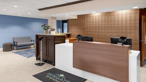Zona de recepción con mostrador de madera, sofá gris, pared azul y felpudo con el texto 'Wingate by Wyndham'.