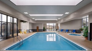 Una piscina interior con paredes de vidrio transparente, un techo con tragaluces y zonas de estar con sillas y mesas.