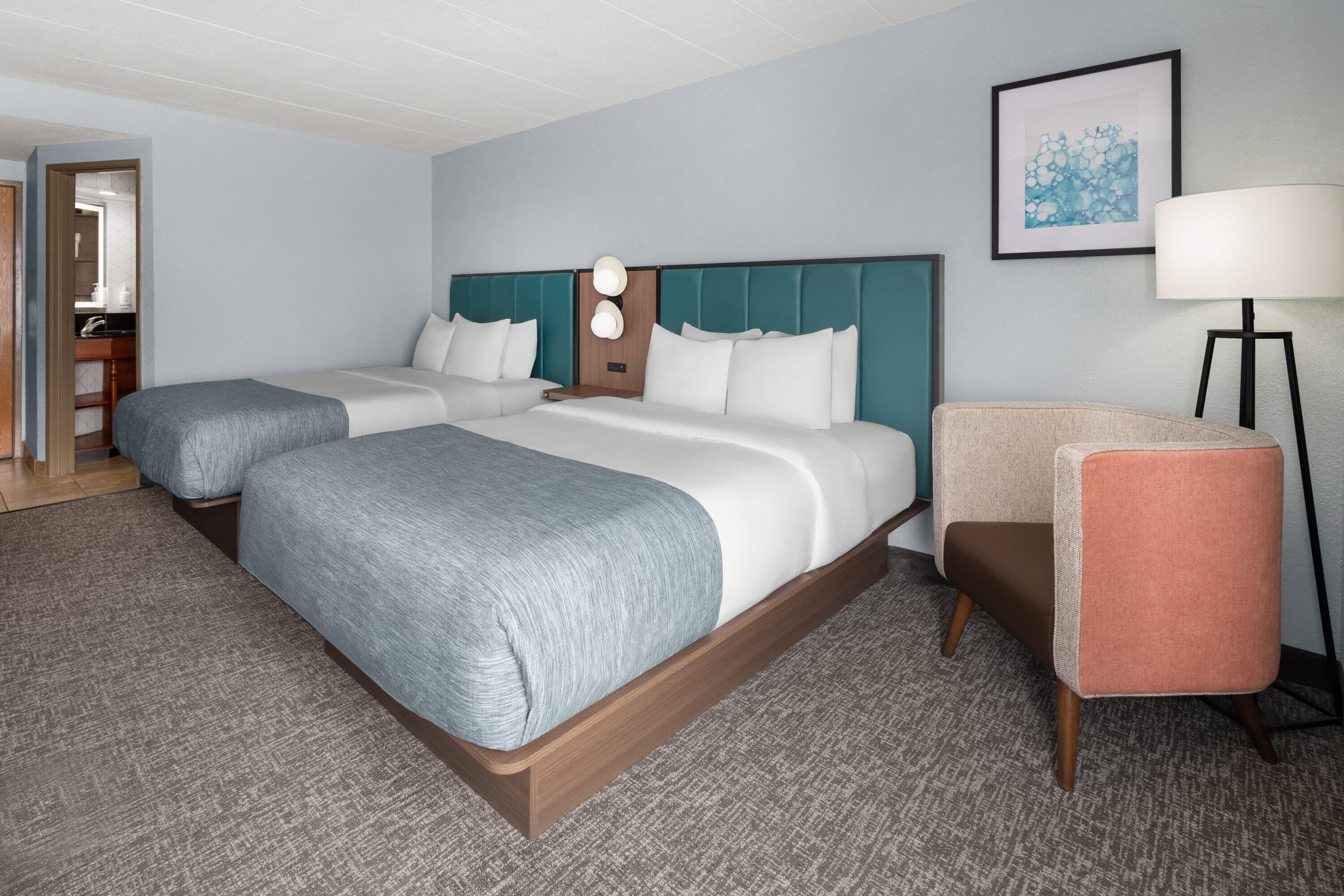 A hotel room with two beds, a desk, a chair, a lamp, and a framed picture on the wall.