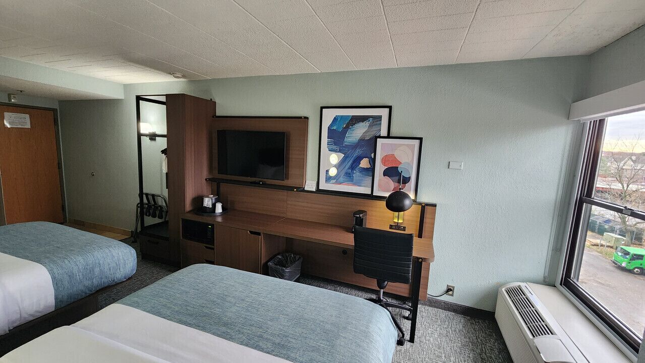 A hotel room with two beds, a desk, a television, and artwork on the wall.