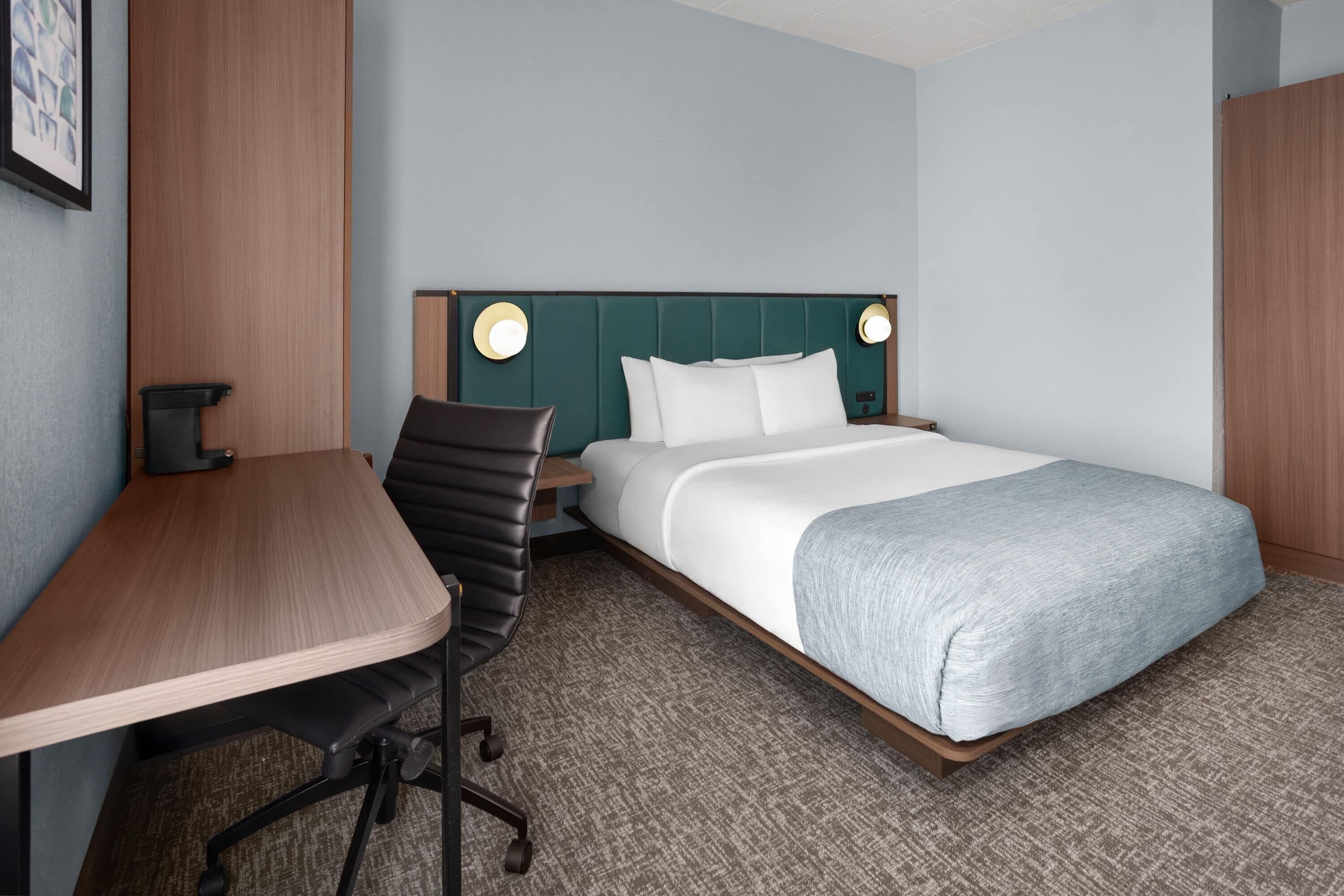 A hotel room with a bed, a desk, a chair, and a wall-mounted lamp.