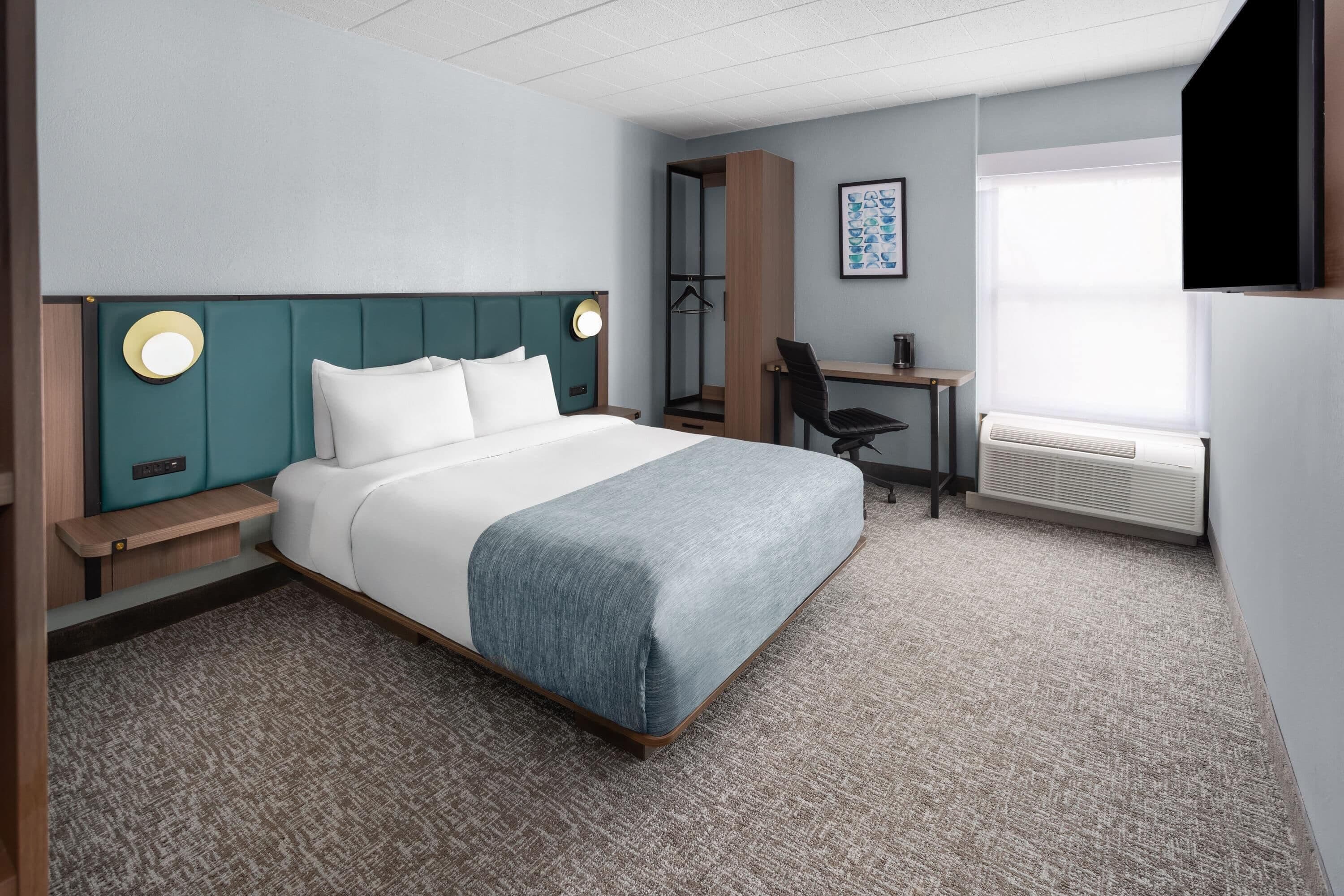 Une chambre d’hôtel avec un lit, un bureau, une chaise, un miroir et une fenêtre.