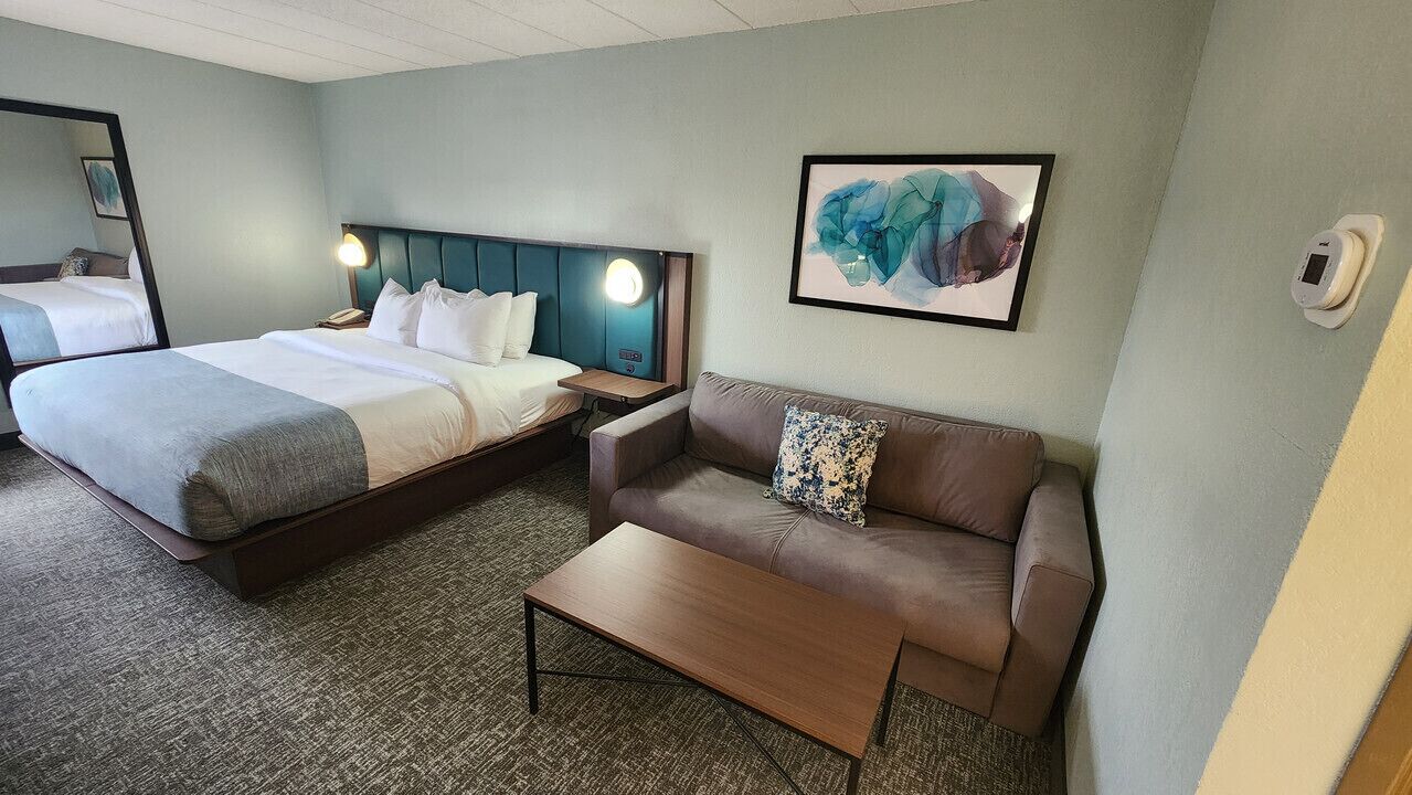 Habitación de hotel con cama, sofá, mesa de centro, mesita de noche, lámpara y una obra de arte enmarcada en la pared.