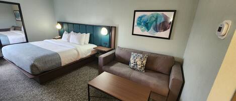 Habitación de hotel con cama, sofá, mesa de centro, mesita de noche, lámpara y una obra de arte enmarcada en la pared.