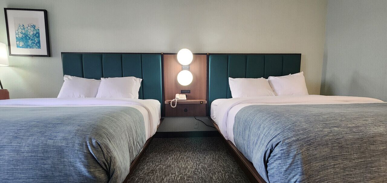 Camera d'albergo con due letti, un comodino con una lampada e un faretto a parete.
