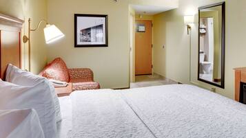 A hotel room with a large bed, a bedside table with a lamp, a chair, a framed picture on the wall, and a door leading to a bathroom.