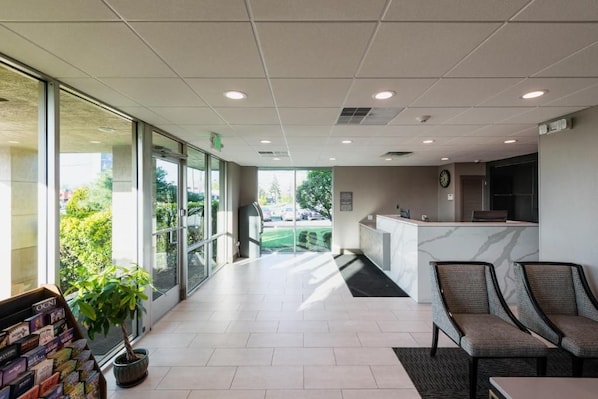 A reception area with a reception desk, chairs, a potted plant, and a view of the outdoors. - The Garden Executive Hotel (South Plainfield)