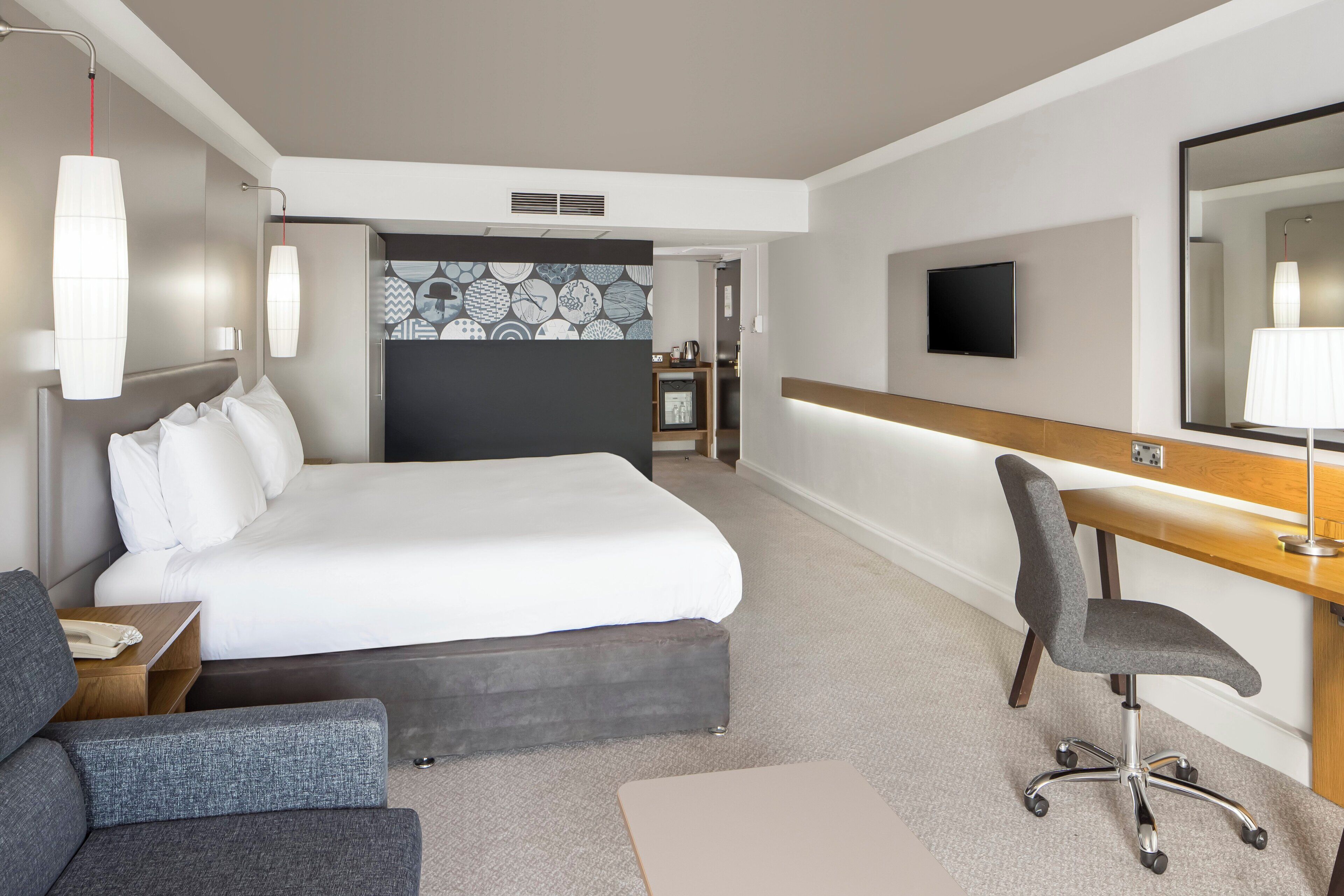 A hotel room with a large bed, a desk with a chair, a television, and a mirror.
