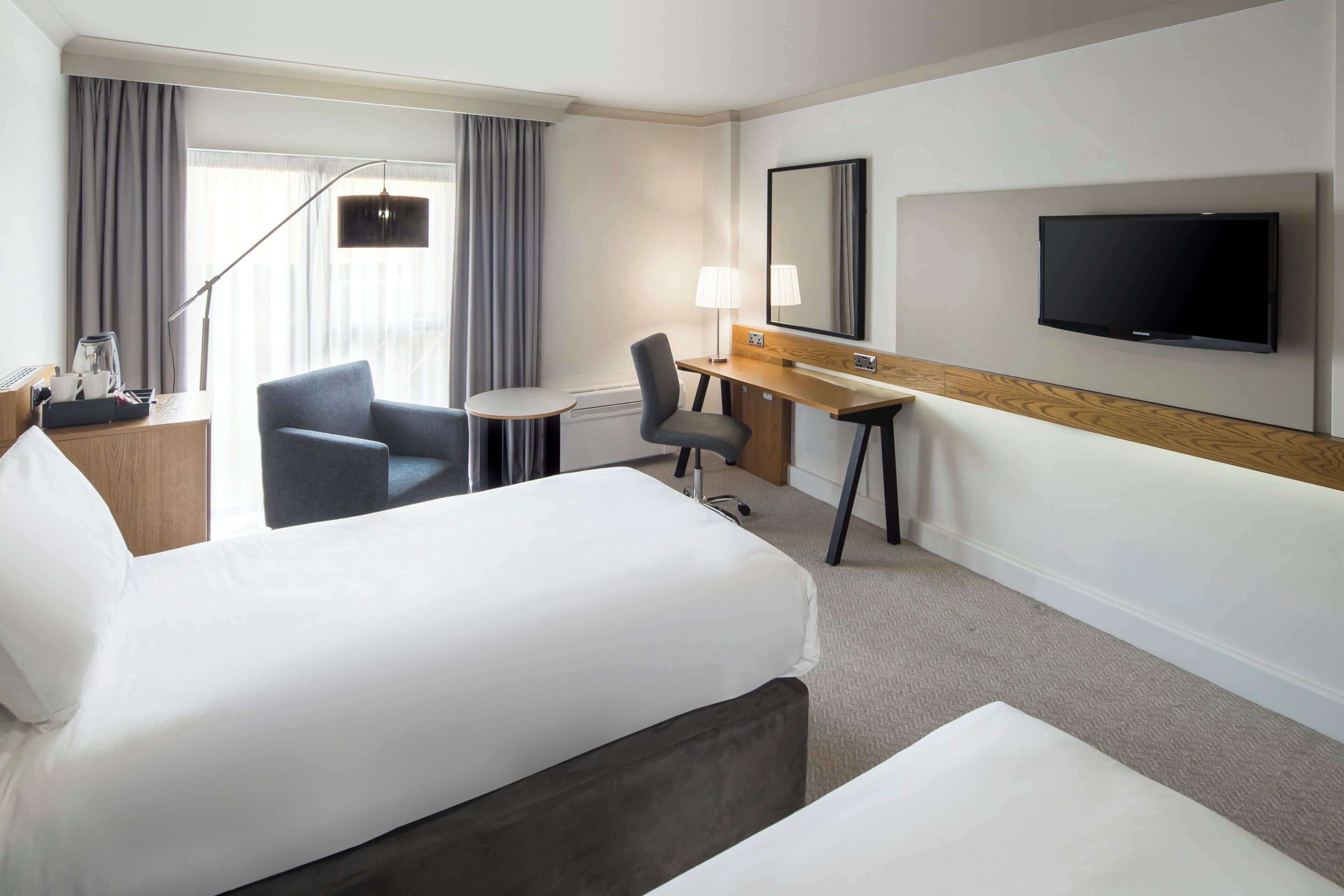A hotel room with two beds, a desk, a chair, a TV, and a window with curtains.