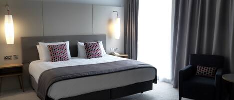 A modern hotel room with a large bed, a bedside table with a lamp, a chair, and a view of the outdoors.