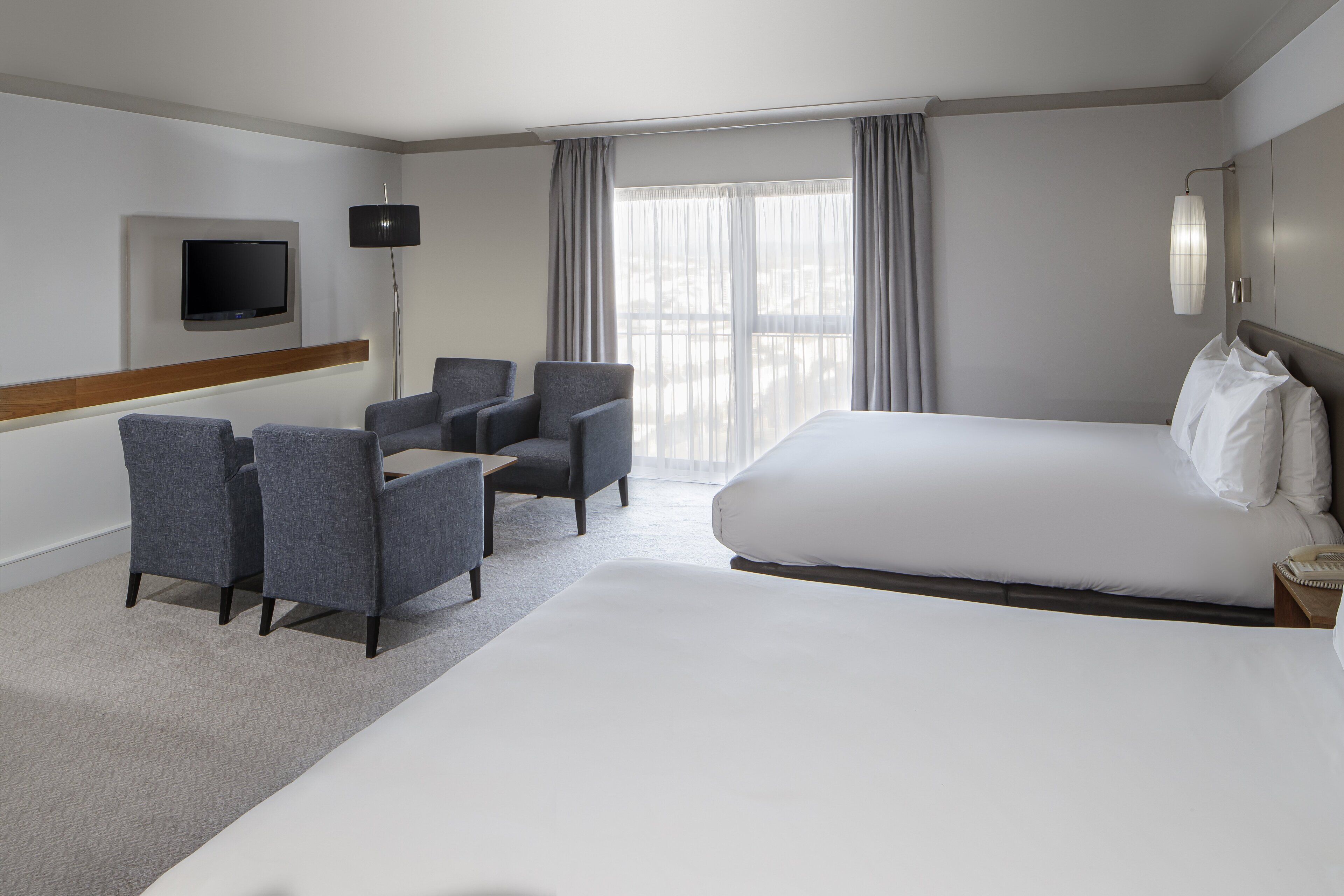 A hotel room with a large bed, two armchairs, a flat-screen TV mounted on the wall, and a large window with curtains.