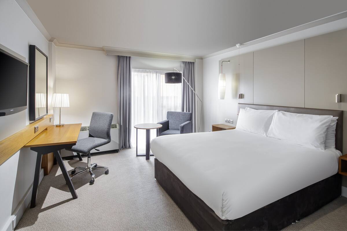 A hotel room with a large bed, a desk with a chair, a small table, and a TV.
