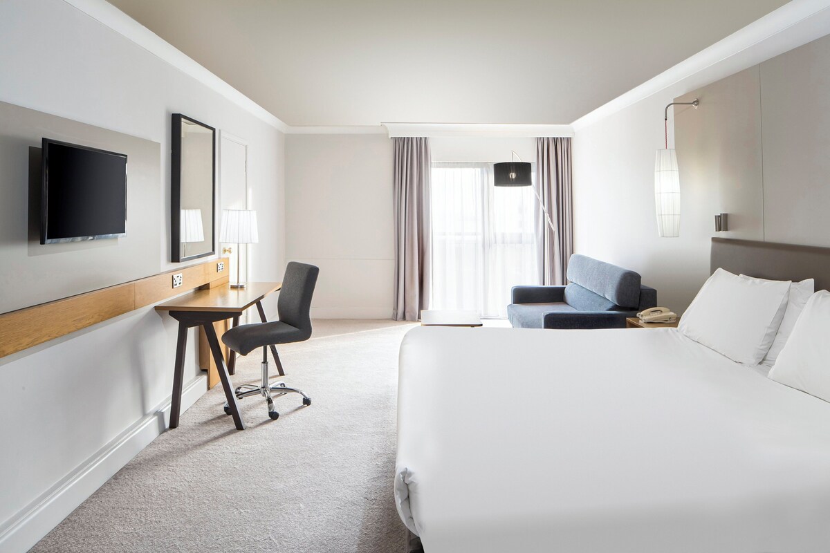 A hotel room with a bed, a desk with a chair, a TV, a lamp, and a window with curtains.