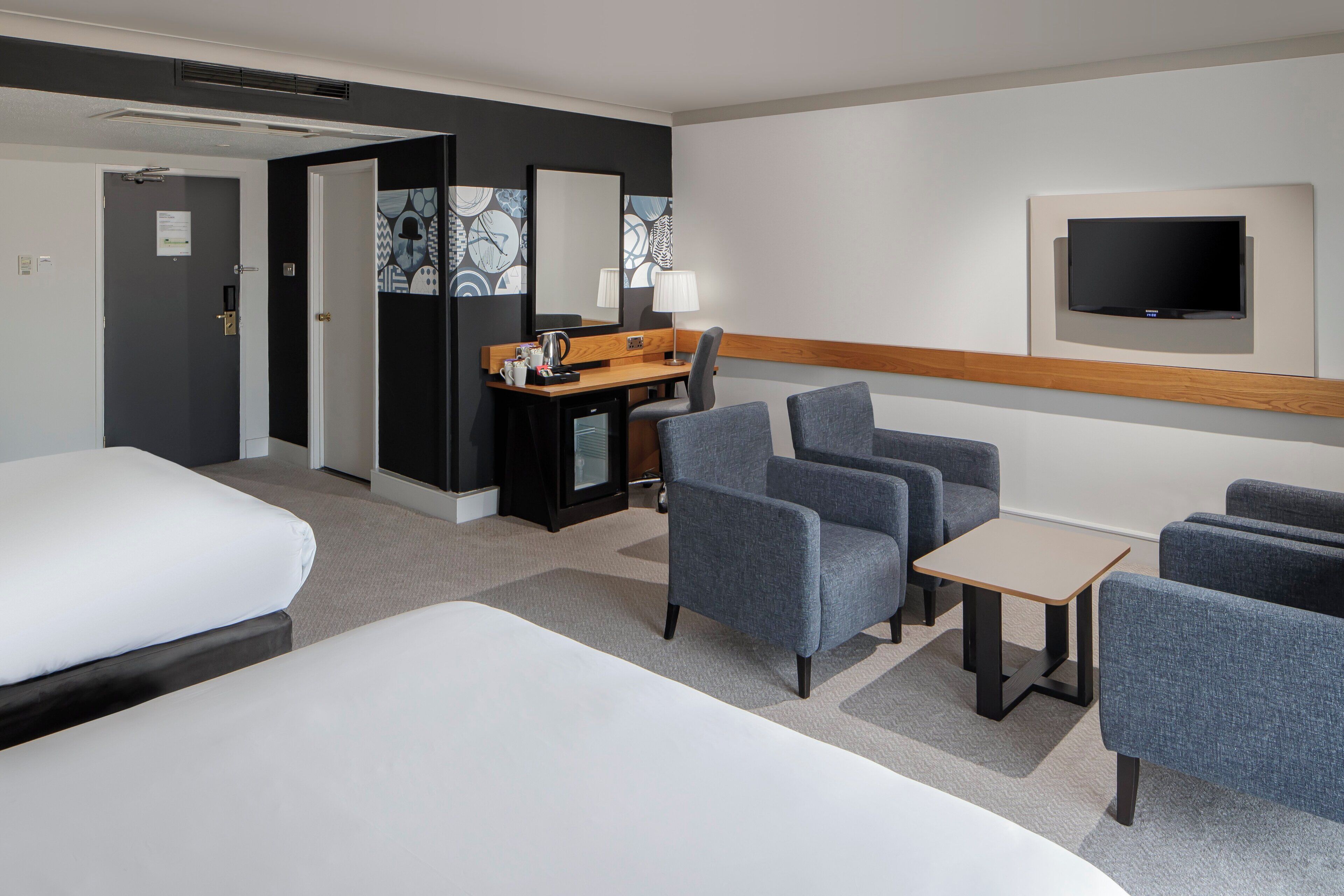 A hotel room with a bed, two armchairs, a small table, a desk with a lamp, and a TV mounted on the wall.