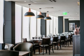 En restaurant med lange borde, mørke stole og hængende lamper.