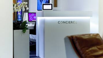Un mostrador de recepción con la palabra 'CONCIERGE' y un monitor de computadora, además de una planta en maceta con flores blancas.