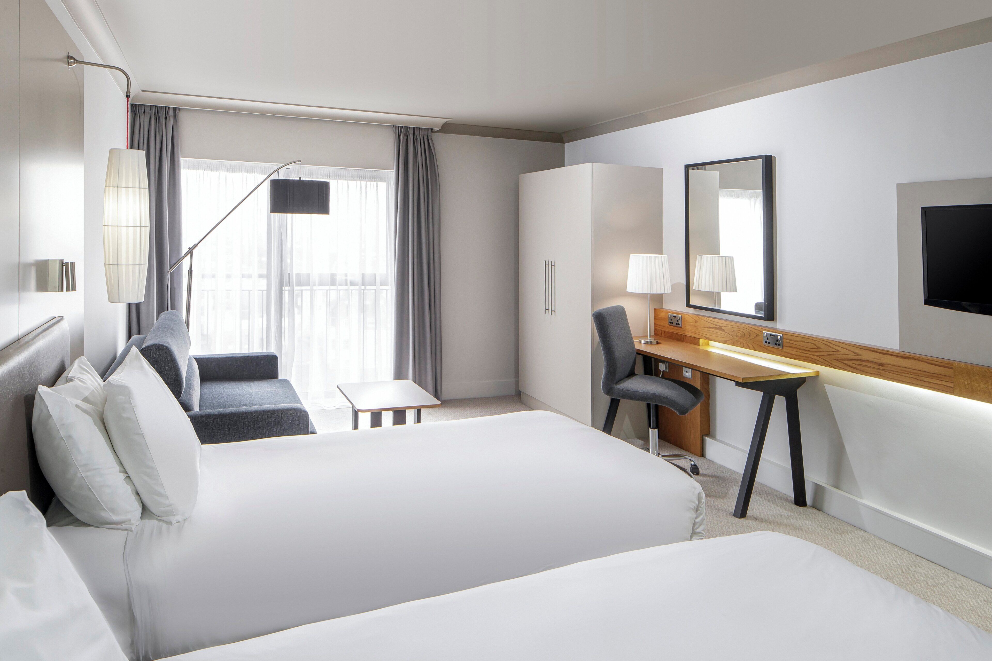 A hotel room with a large bed, a desk, a chair, a TV, and a window with curtains.
