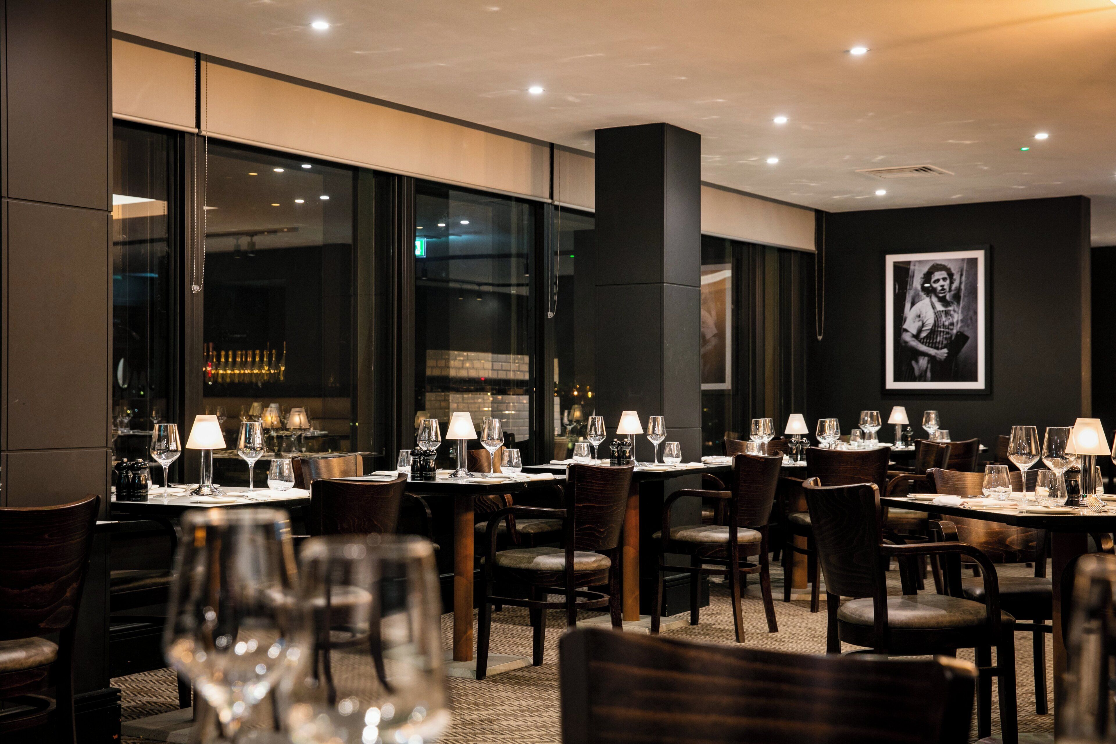 Interior de un restaurante con mesas dispuestas para comer, sillas y una zona de bar visible a través de la ventana.