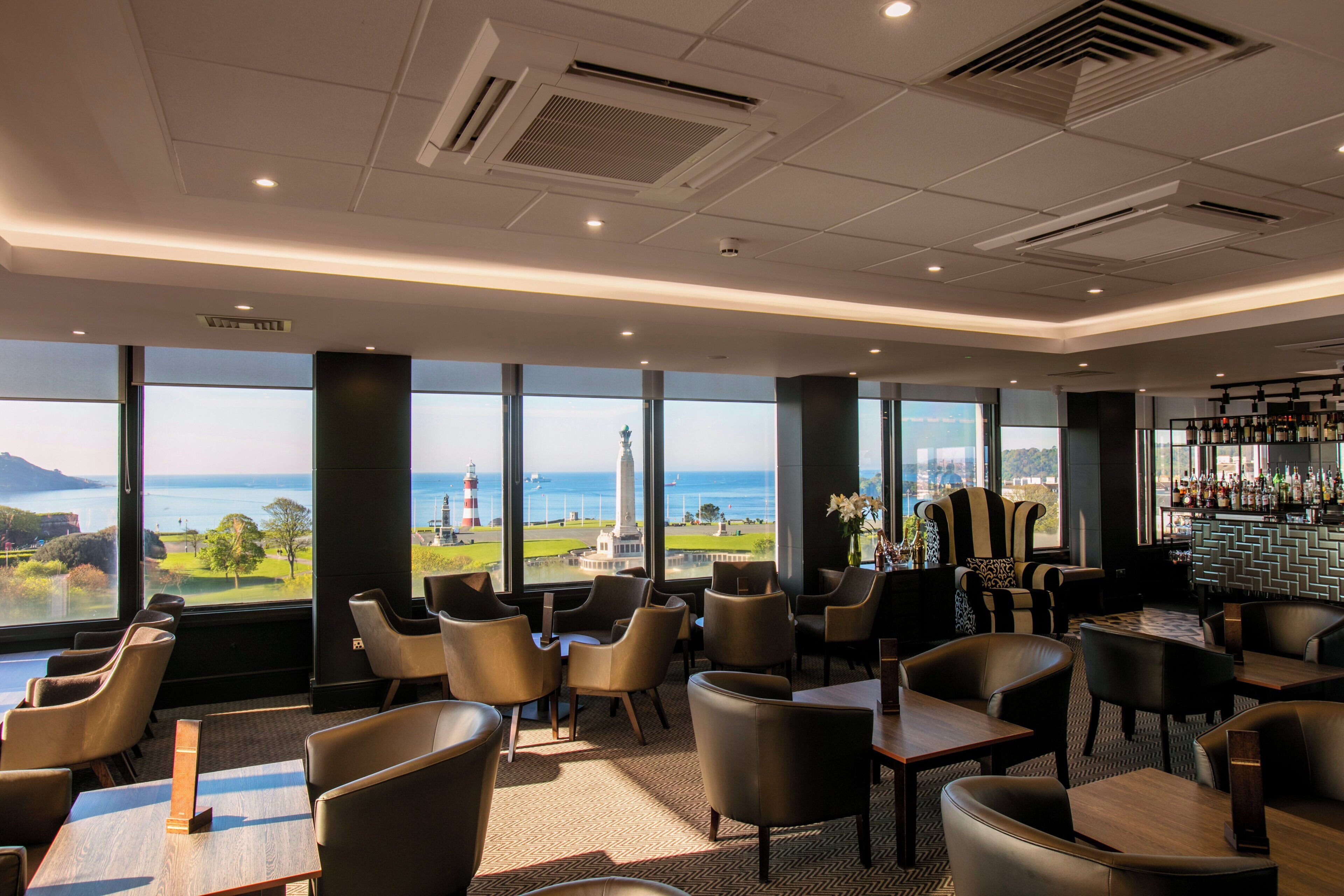 A hotel bar with multiple seating areas, large windows offering a view of the sea and a lighthouse, and a bar stocked with bottles.