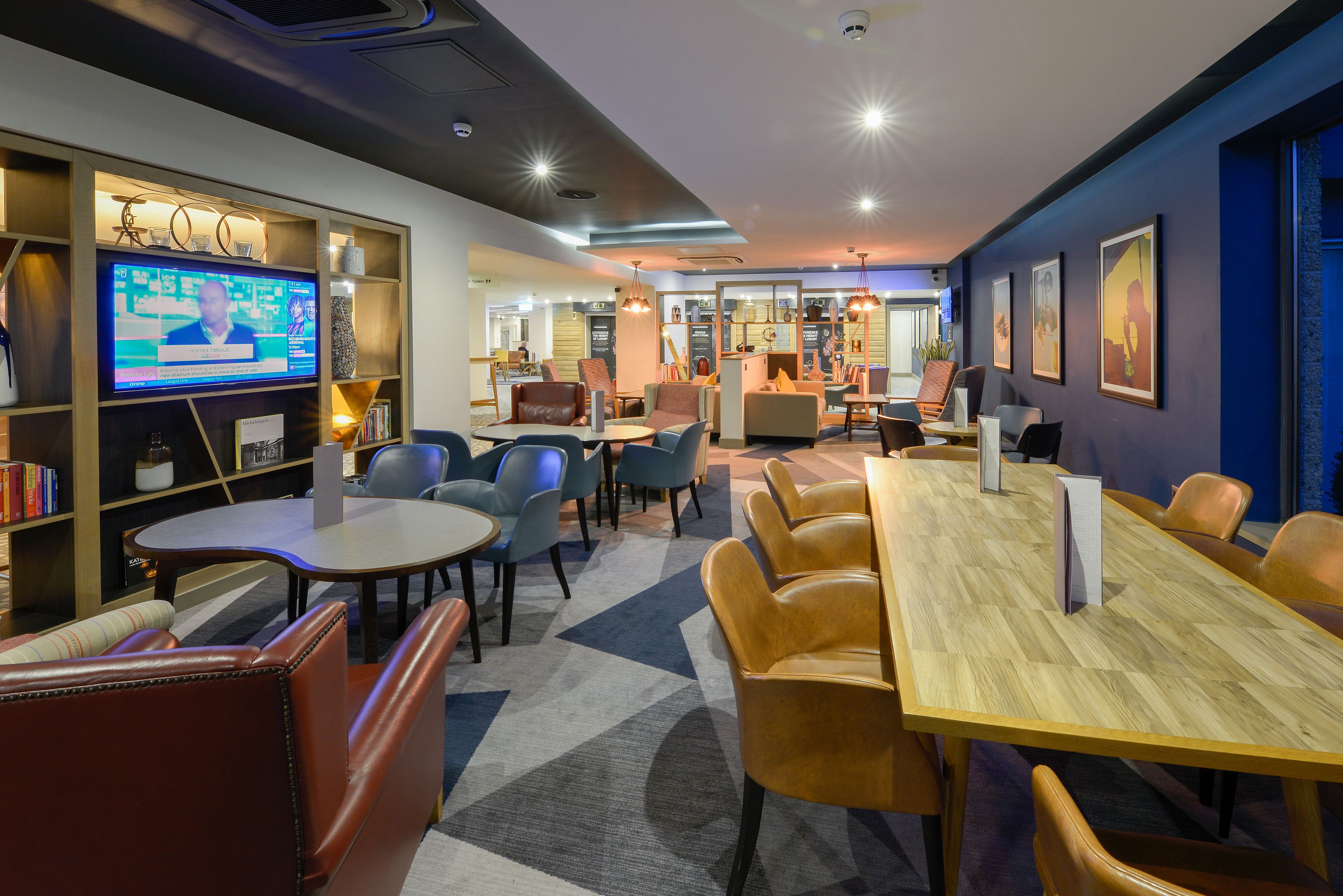 A modern hotel lobby with a large TV screen, comfortable seating, and a mix of tables and chairs.