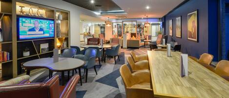 A modern hotel lobby with a large TV screen, comfortable seating, and a mix of tables and chairs.