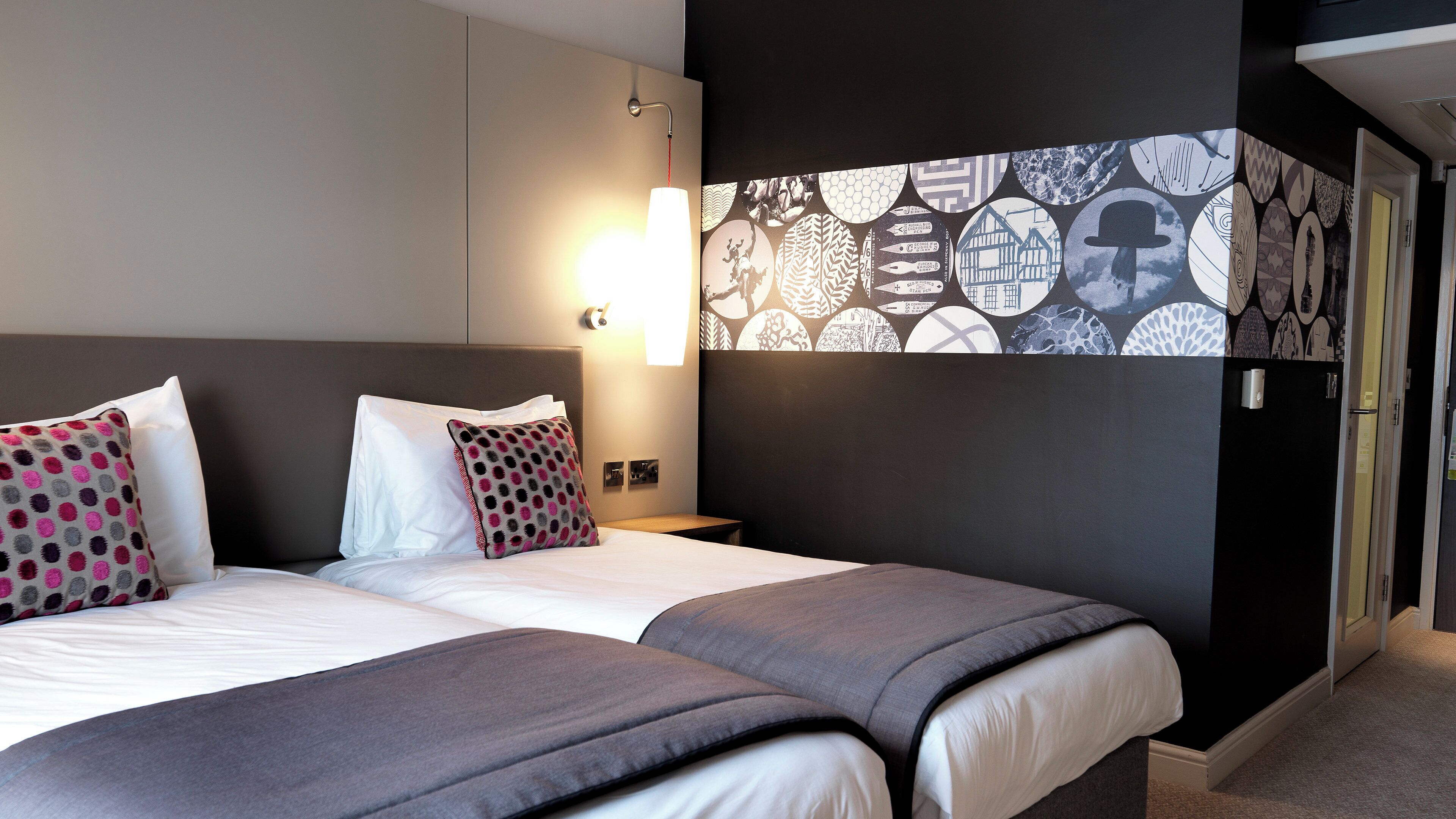 A hotel room with a double bed, two pillows, a bedside table, a wall-mounted light, and a patterned wall decoration.