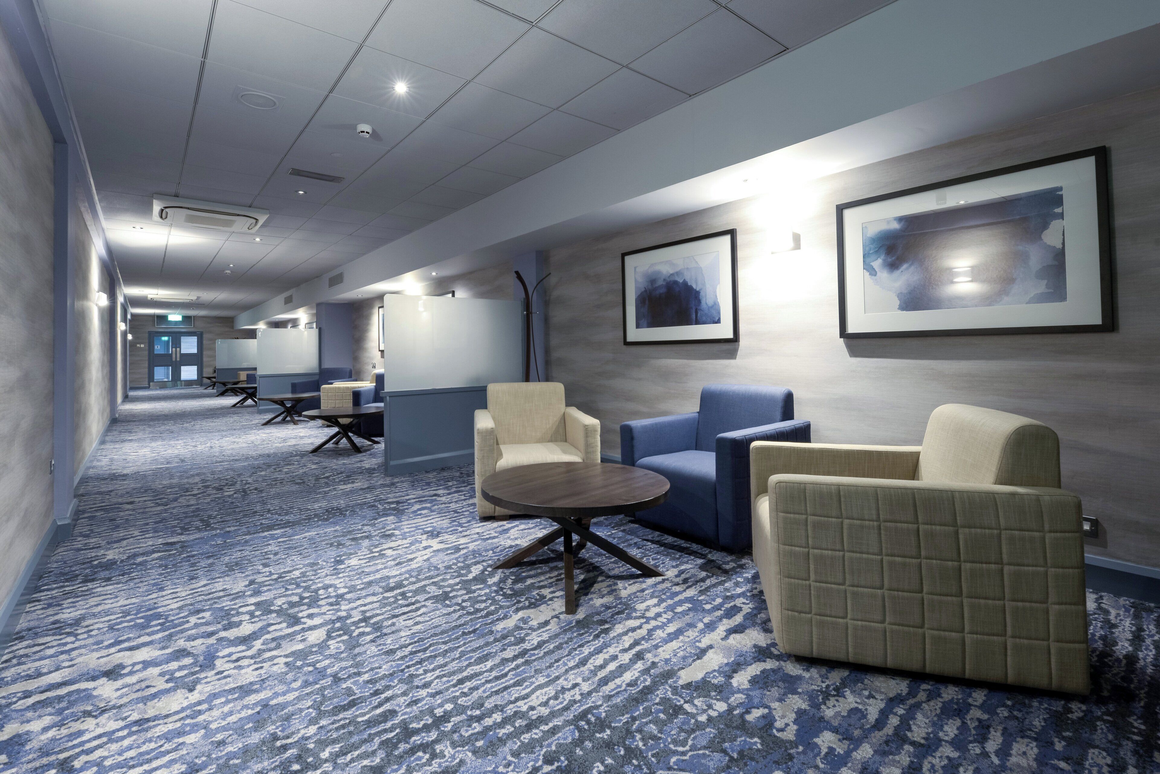 A hotel corridor with seating areas, a patterned carpet, and framed artwork on the walls.
