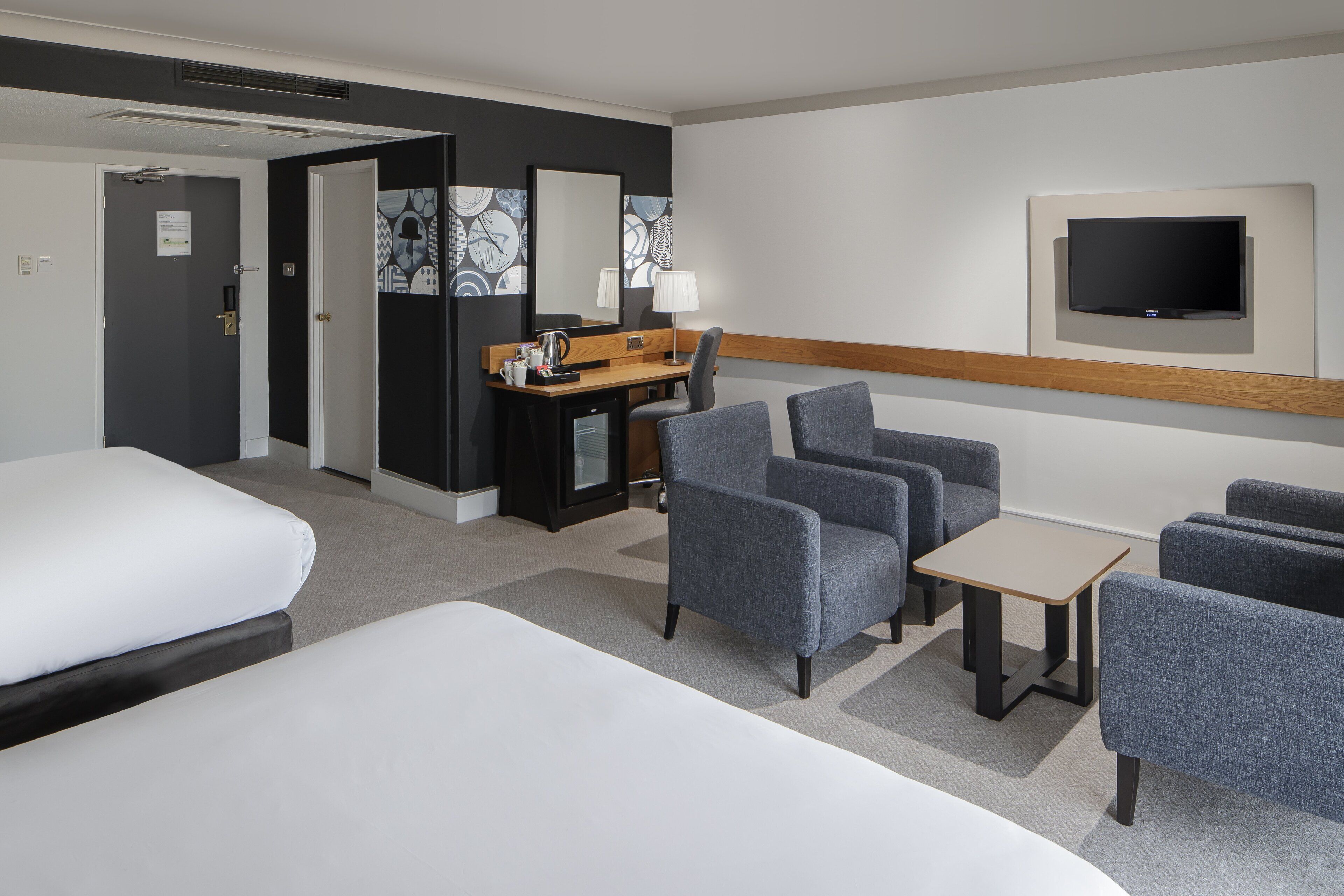 A hotel room with a bed, a desk with a lamp, a TV mounted on the wall, and several chairs.