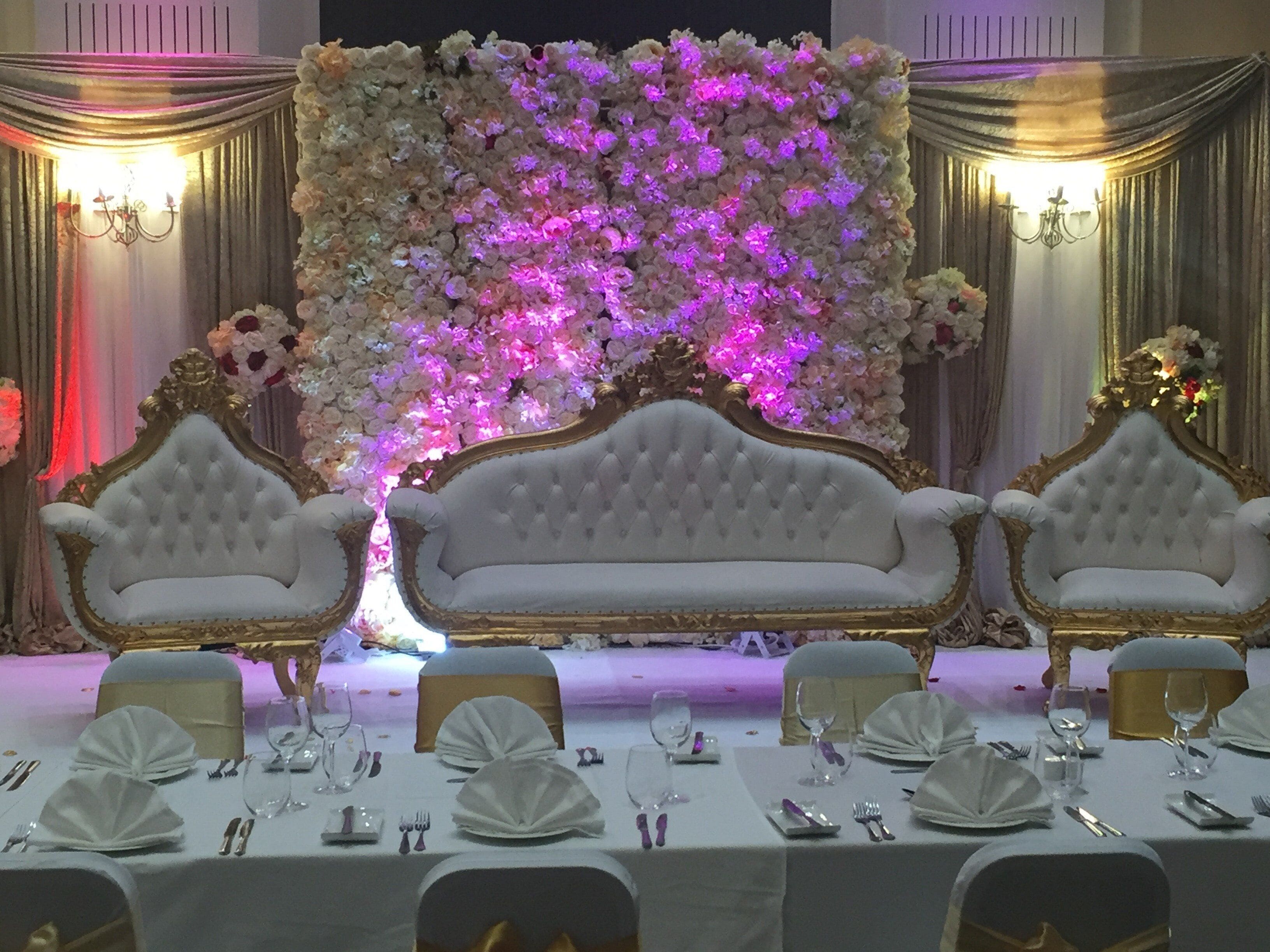 A banquet hall with a central stage, ornate white and gold chairs, and a floral backdrop.