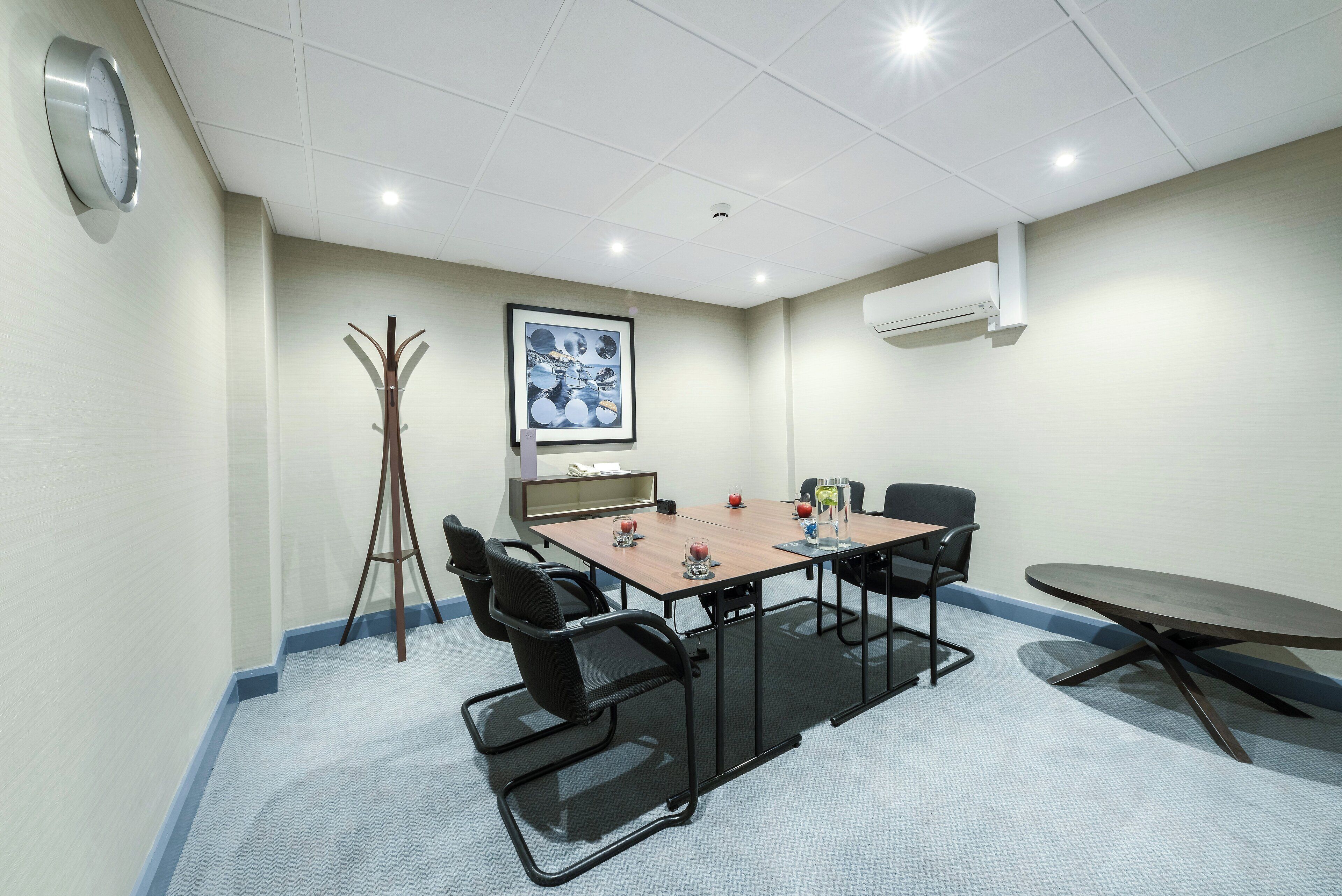 Une salle de conférence avec une table rectangulaire, six chaises, une horloge, un porte-manteau et un tableau encadré au mur.