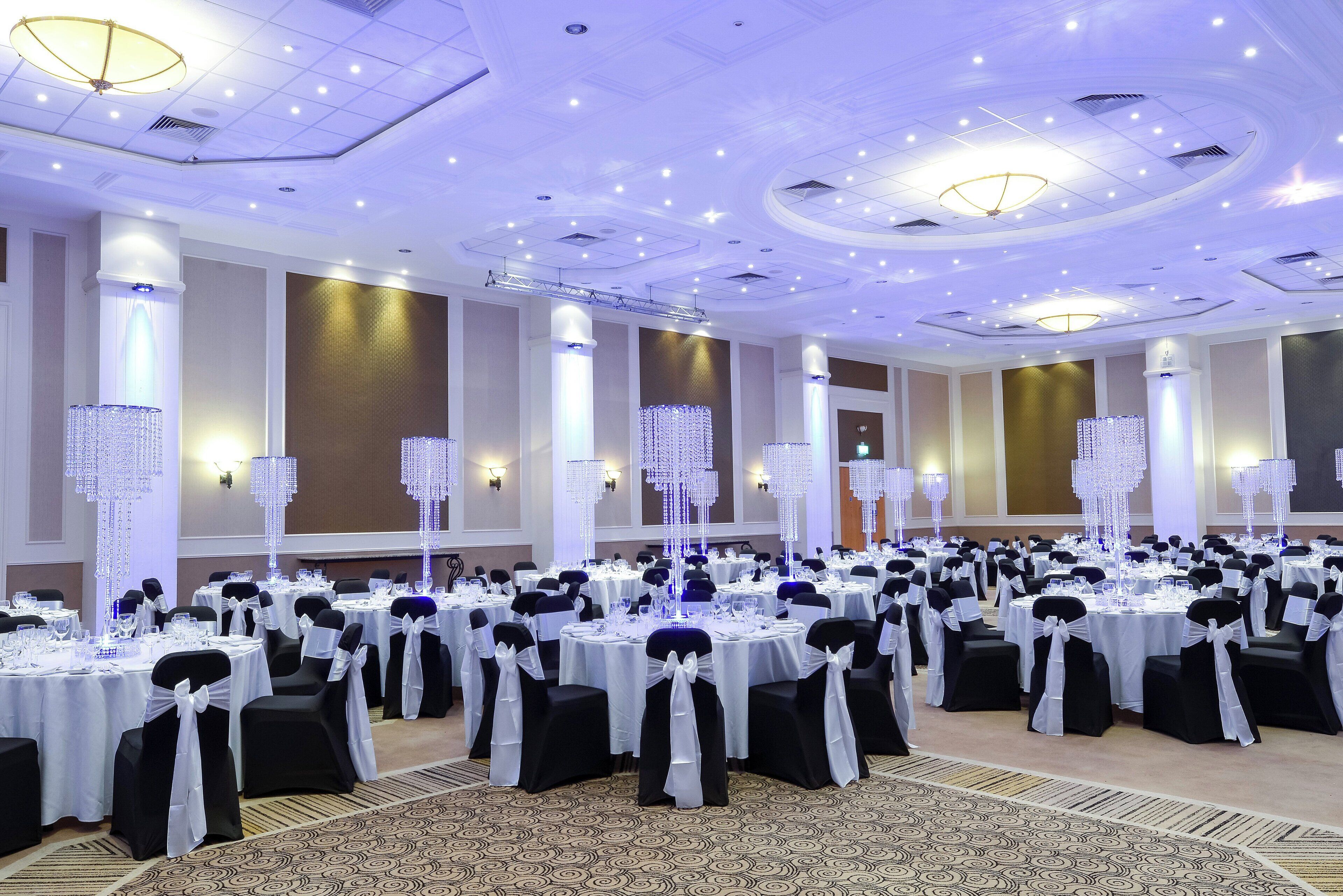 A banquet hall with round tables, black and white chairs, and decorative crystal chandeliers.