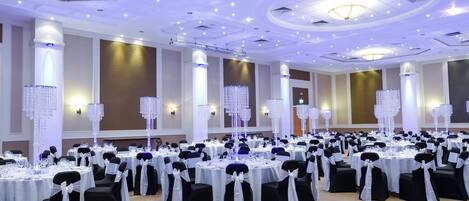 A banquet hall with round tables, black and white chairs, and decorative crystal chandeliers.