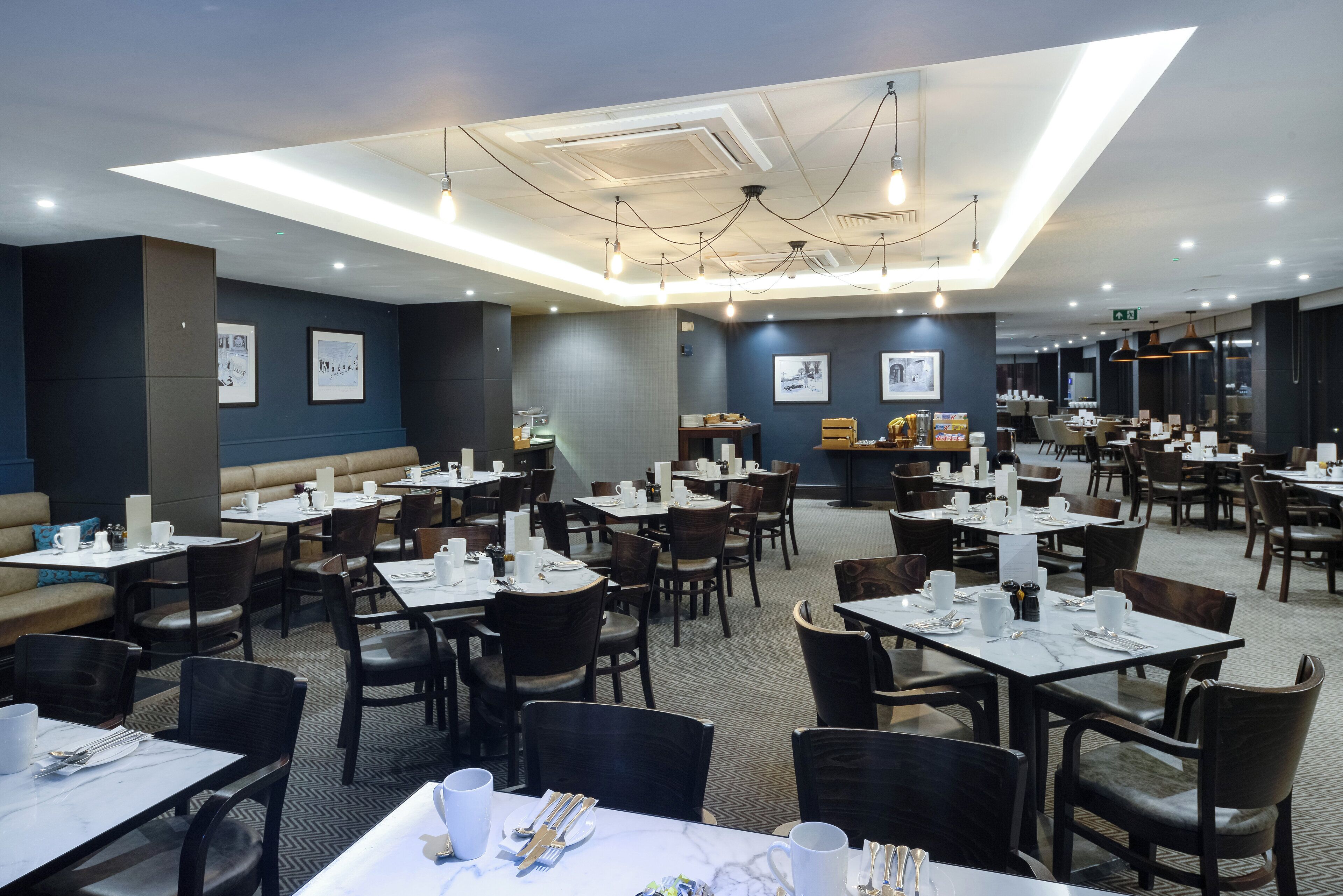 A hotel dining area with multiple tables and chairs, ceiling lights, and framed pictures on the walls.