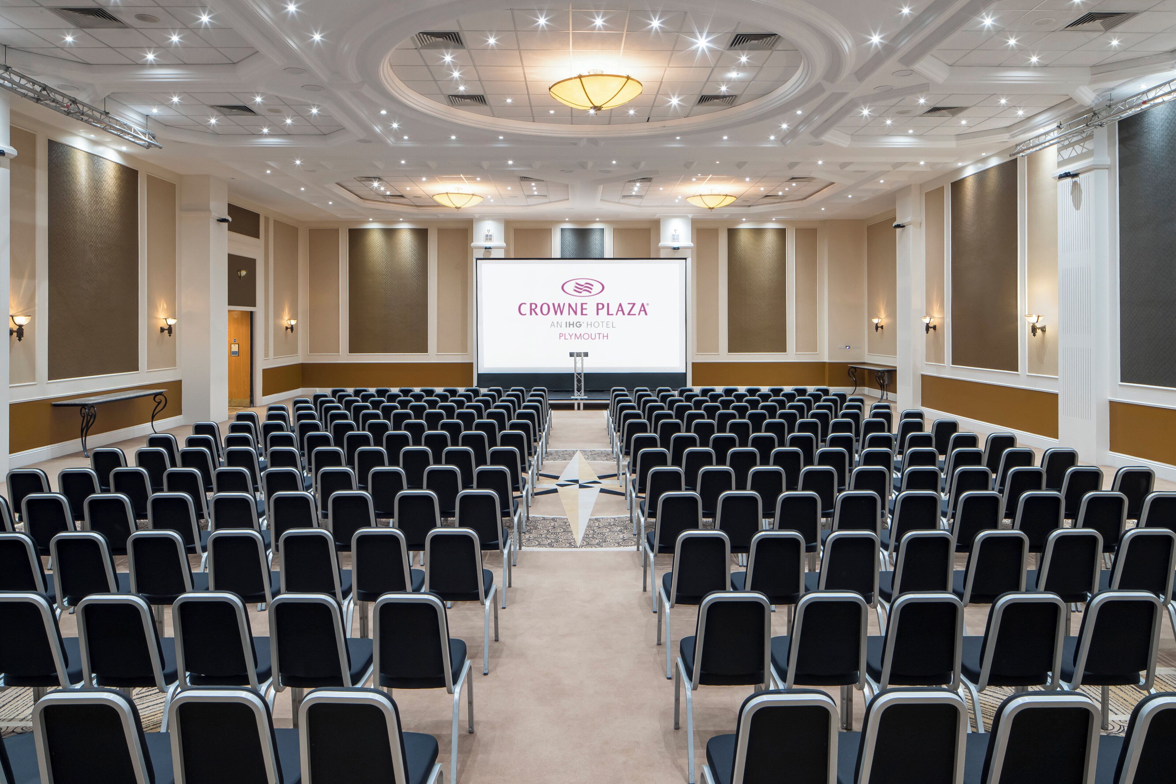 Un salón de conferencias con filas de sillas que dan a una pantalla de proyección que muestra 'CROWNE PLAZA AN IHG HOTEL PLYMOUTH'.