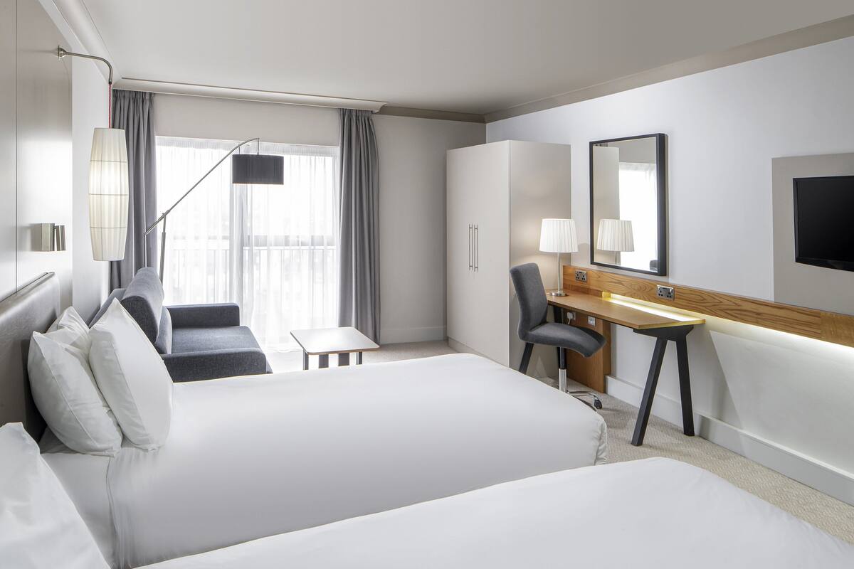 A hotel room with two beds, a desk, a chair, a TV, and a large window with curtains.